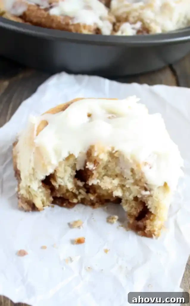 A 1 hour cinnamon roll with a bite missing on a piece of paper. A pan of cinnamon rolls rests in the background. 
