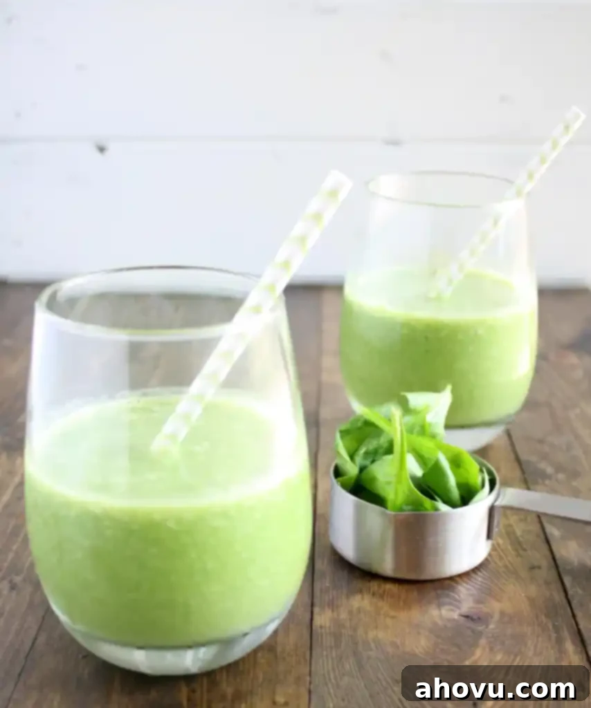 Side view of two spinach fruit smoothies with straws. A measuring cup of spinach rests between the glasses. 