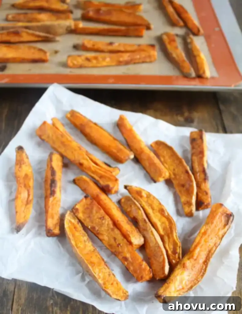 Oven baked sweet potato fries on a square of parchment paper. More fries are on a baking sheet in the background. 