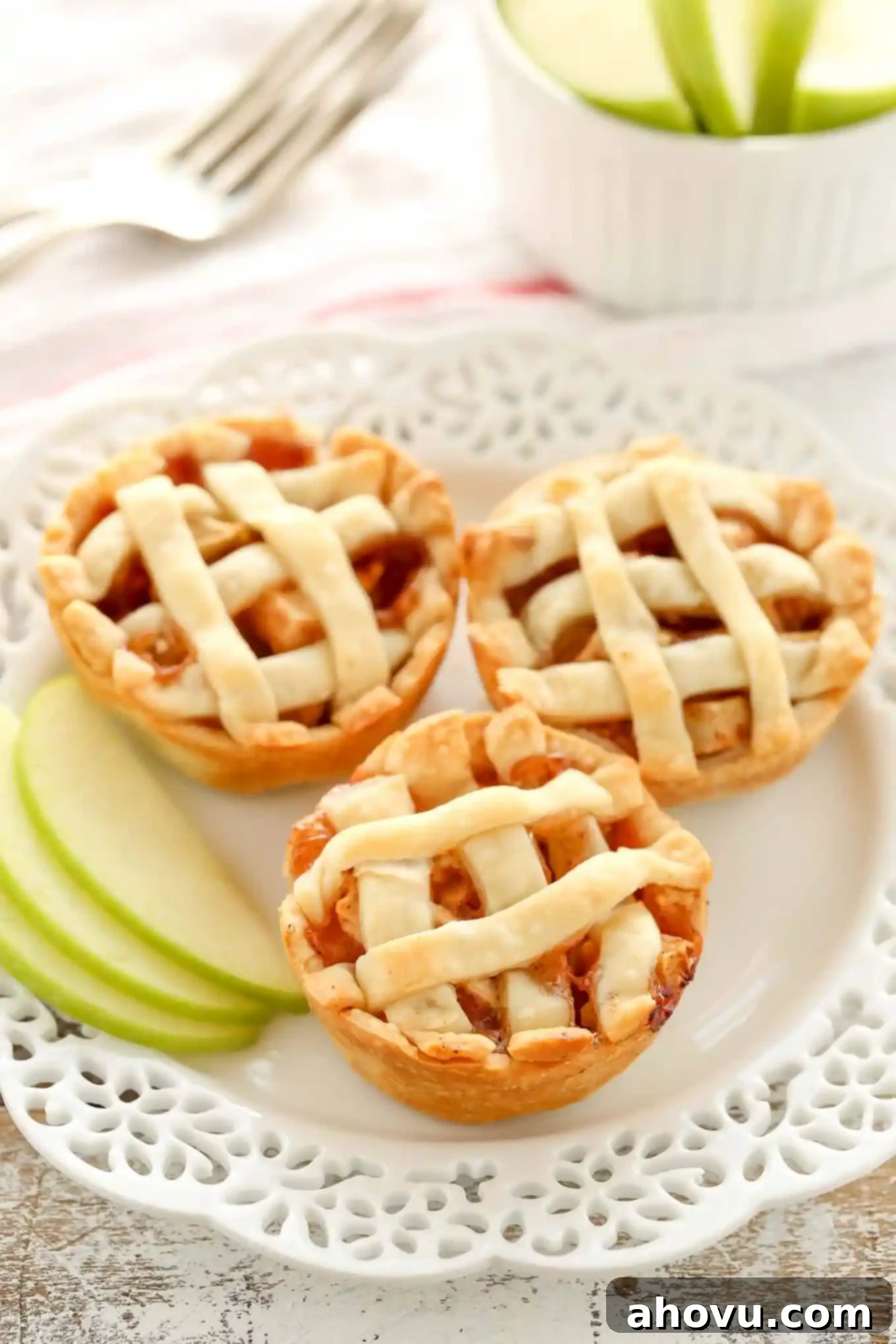 Three mini apple pies on a decorative white plate. A small container of sliced apples and a fork are in the background.