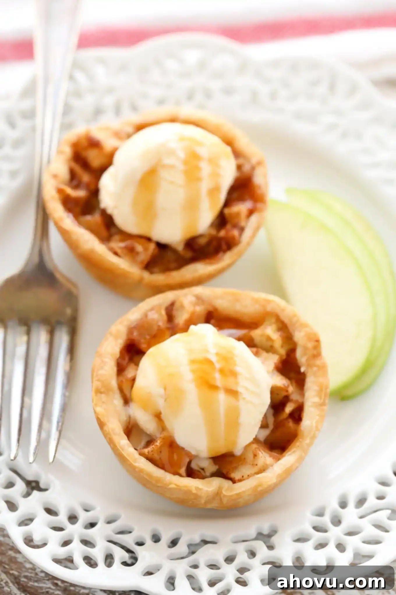 Two mini apple pies topped with a small scoop of ice cream and a drizzle of salted caramel sauce. A fork and a few slices of apples lay on the plate next to the pies.