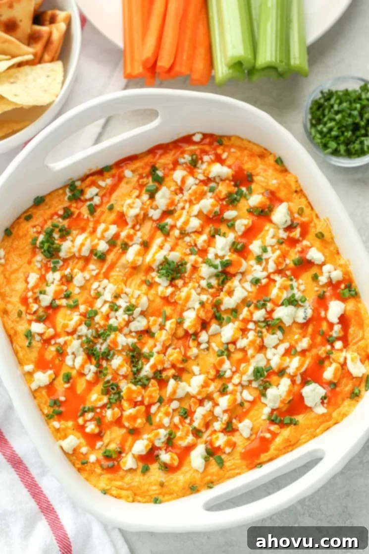 Ultimate Buffalo Chicken Dip 2 A white baking dish filled with creamy buffalo chicken dip topped with blue cheese, chives, and buffalo sauce. A bowl of chips, a plate of celery and carrot sticks and small bowl of chives around the dish.