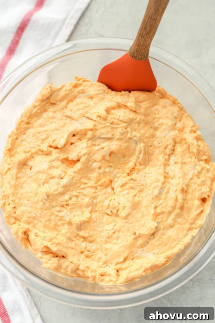 Ultimate Buffalo Chicken Dip 3 A clear glass bowl filled with creamy buffalo chicken dip with a rubber spatula leaning on the side. There is a white napkin in the background.