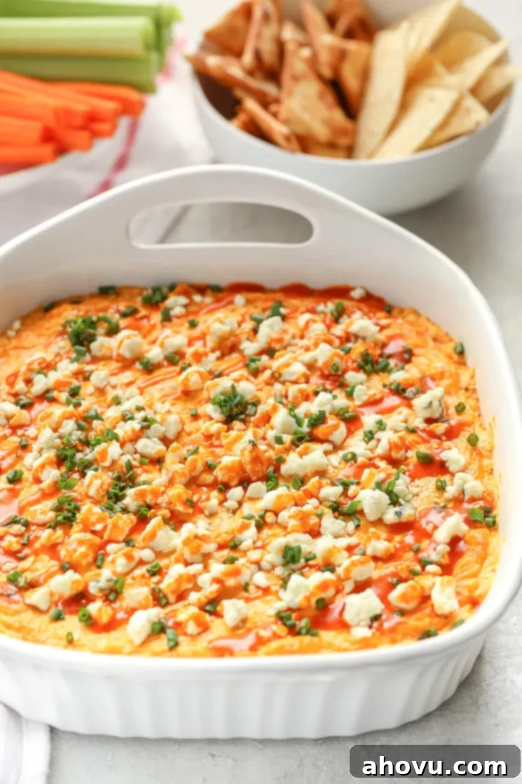 Ultimate Buffalo Chicken Dip 5 A white baking dish filled with creamy buffalo chicken dip topped with blue cheese, chives, and buffalo sauce. A bowl of chips and a plate with celery and carrot sticks in the background.