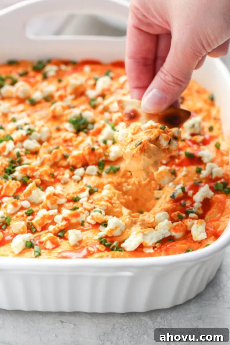 Ultimate Buffalo Chicken Dip 6 A white baking dish filled with creamy buffalo chicken dip topped with blue cheese, chives, and buffalo sauce. A hand holding a pita chip dipping into the buffalo chicken dip.