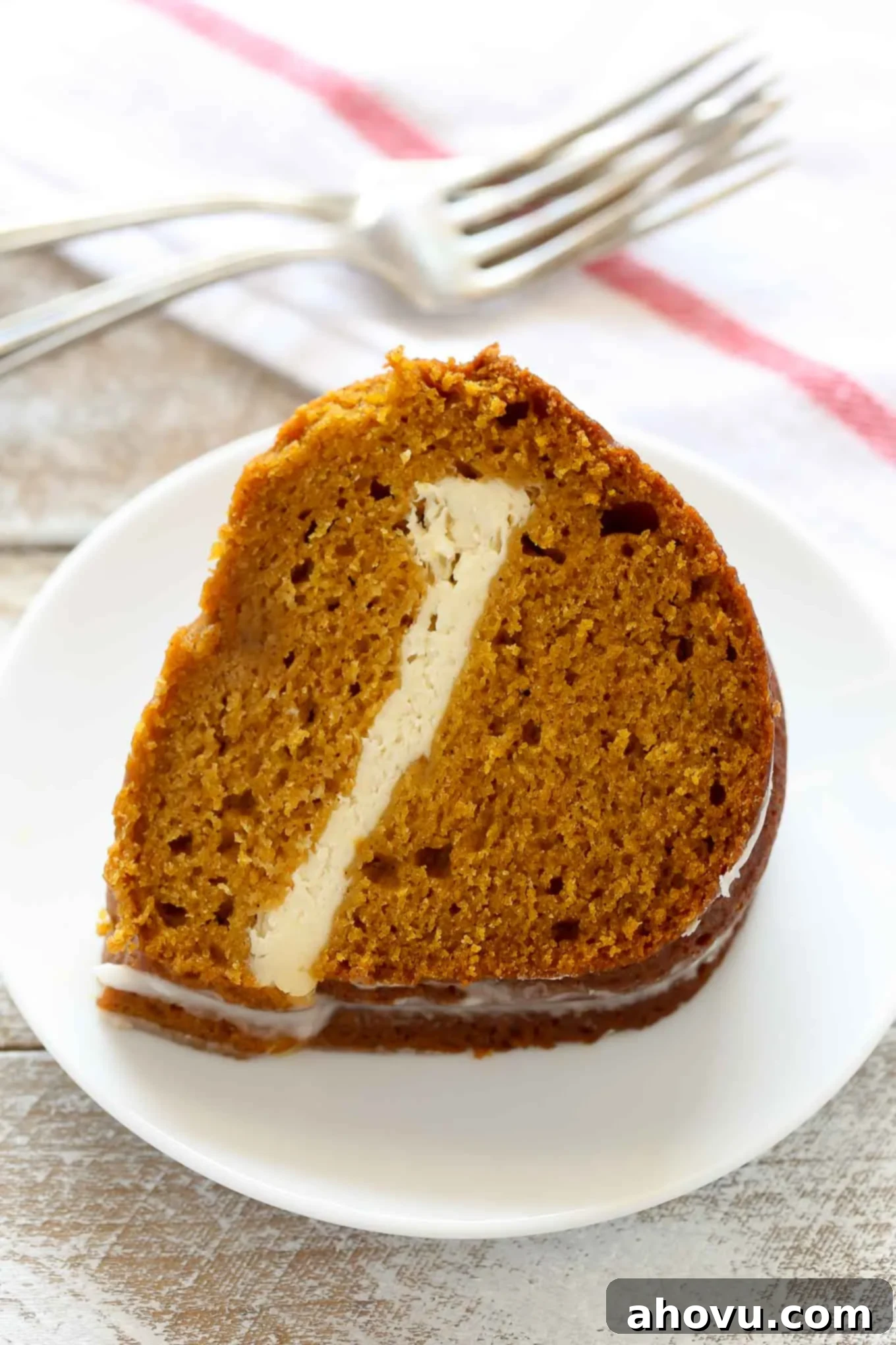 Pumpkin Swirl Cream Cheese Bundt 5 A perfectly portioned slice of pumpkin cream cheese cake, showcasing its rich layers, sits on a white plate, with two forks and a tea towel softly blurred in the background, ready for enjoyment.