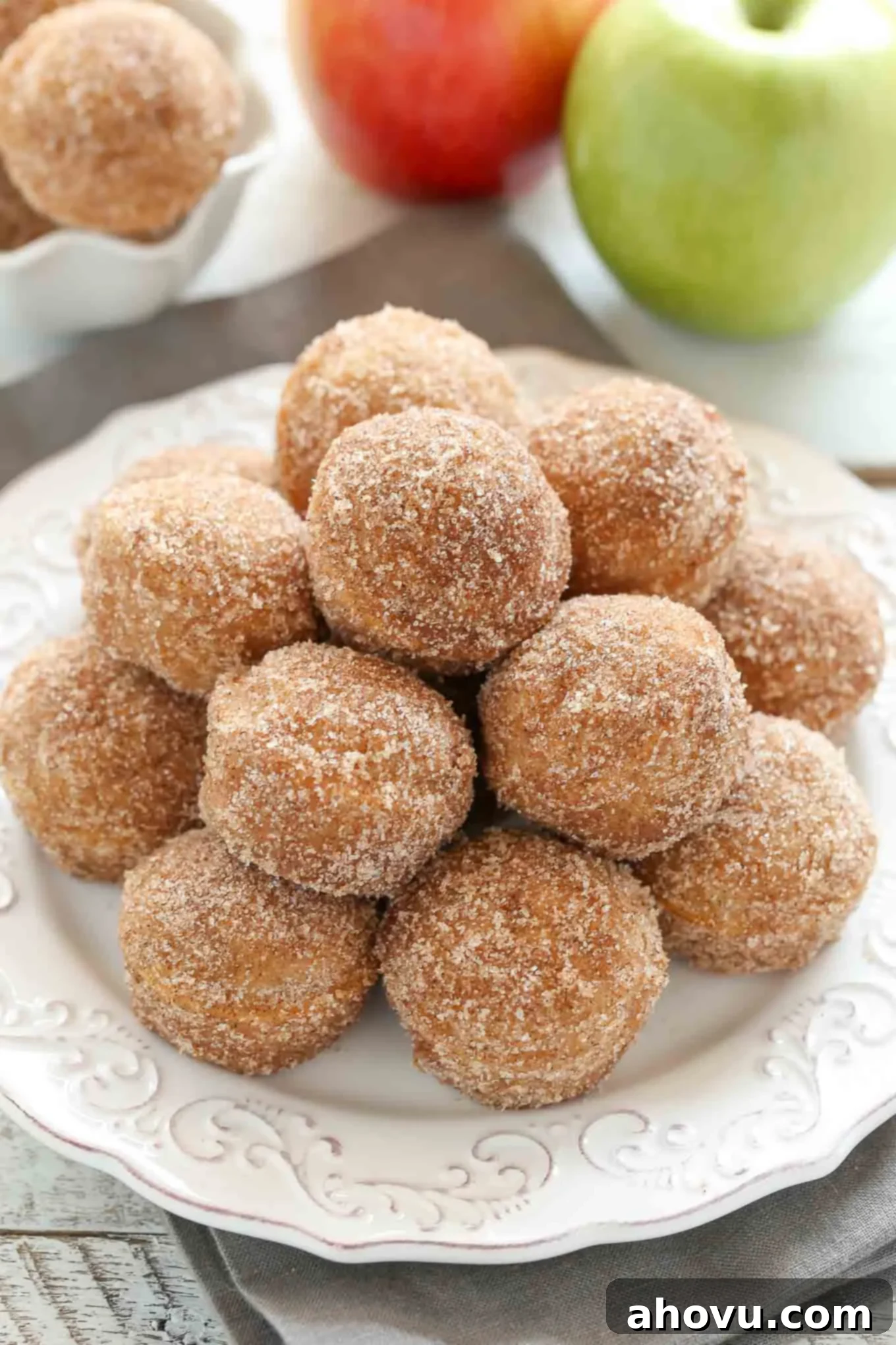 Oven-Fresh Apple Cider Donut Bites 2 A white plate piled high with golden-brown apple cider donut holes. Two fresh red apples are gently placed in the background, hinting at the key ingredient.