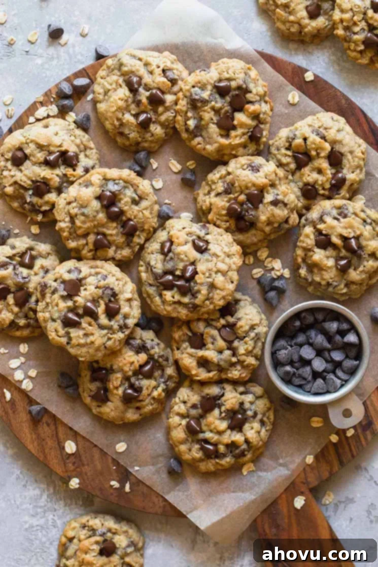 A wooden tray with freshly baked soft and chewy oatmeal chocolate chip cookies on parchment paper, surrounded by scattered raw oats and chocolate chips, highlighting their delicious ingredients and homemade appeal.