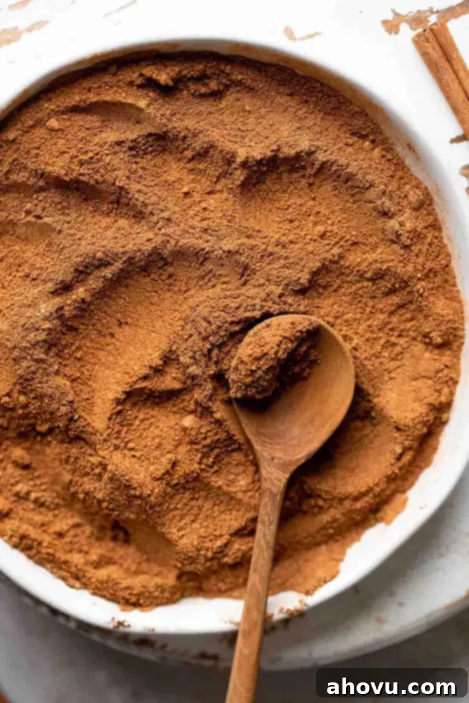 Pumpkin pie spice mixed together in a white dish. A wooden spoon rests on top of the spices and a cinnamon stick rests on the side.
