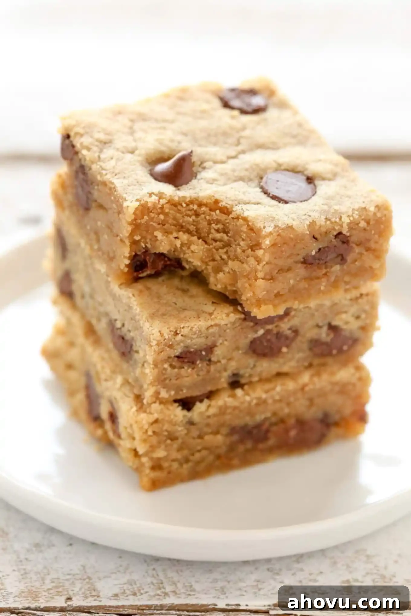 A stack of three peanut butter chocolate chip bars on a white plate. The top bar has a bite missing. 