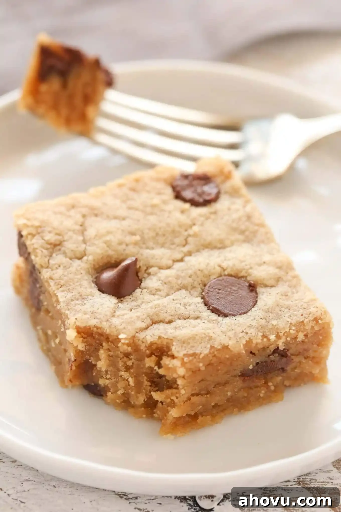 A peanut butter chocolate chip cookie bar on a white plate with a fork. A bite has been taken from the bar. 