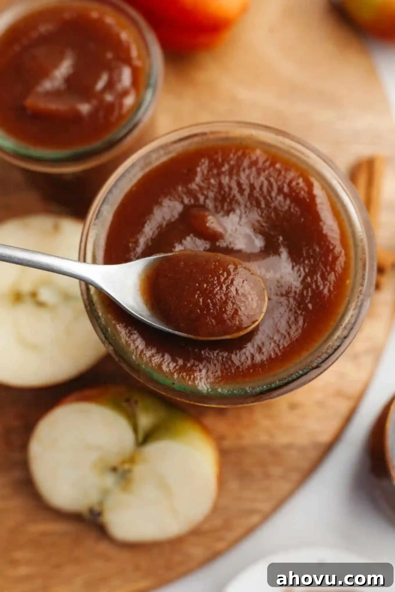 Homemade Spiced Apple Butter 2 A glass jar filled with rich, brown apple butter, with a spoon dipping into it. The background shows autumn leaves and spices, highlighting the fall theme of this slow cooker apple butter.