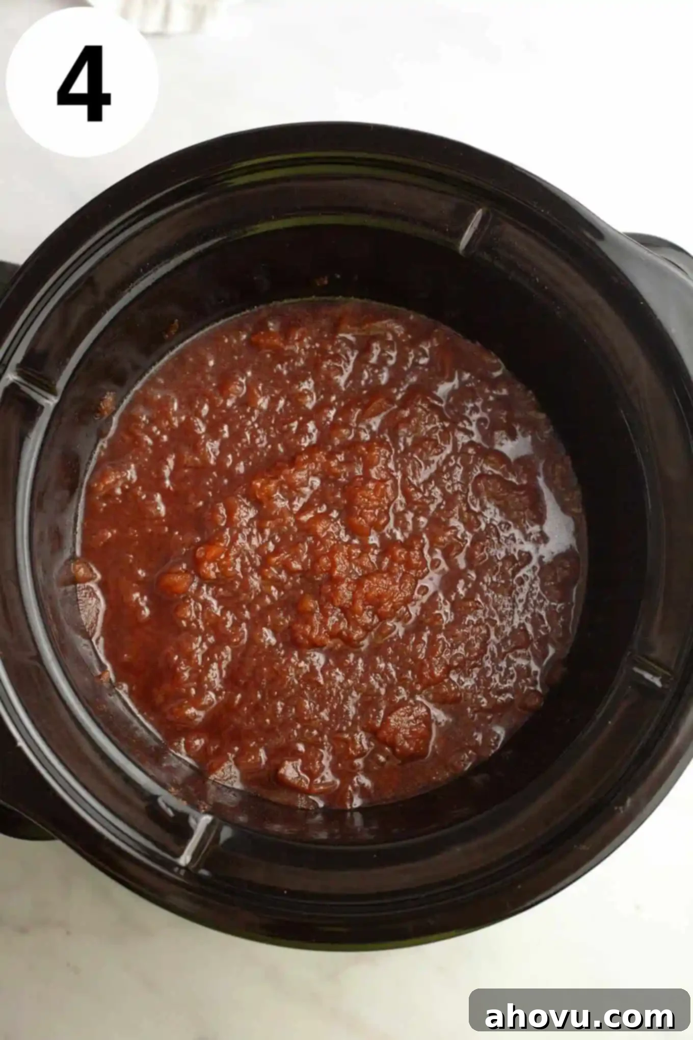 Homemade Spiced Apple Butter 7 An overhead view of chunky, cooked apples in a slow cooker, mashed with a potato masher, illustrating an intermediate step in making apple butter.