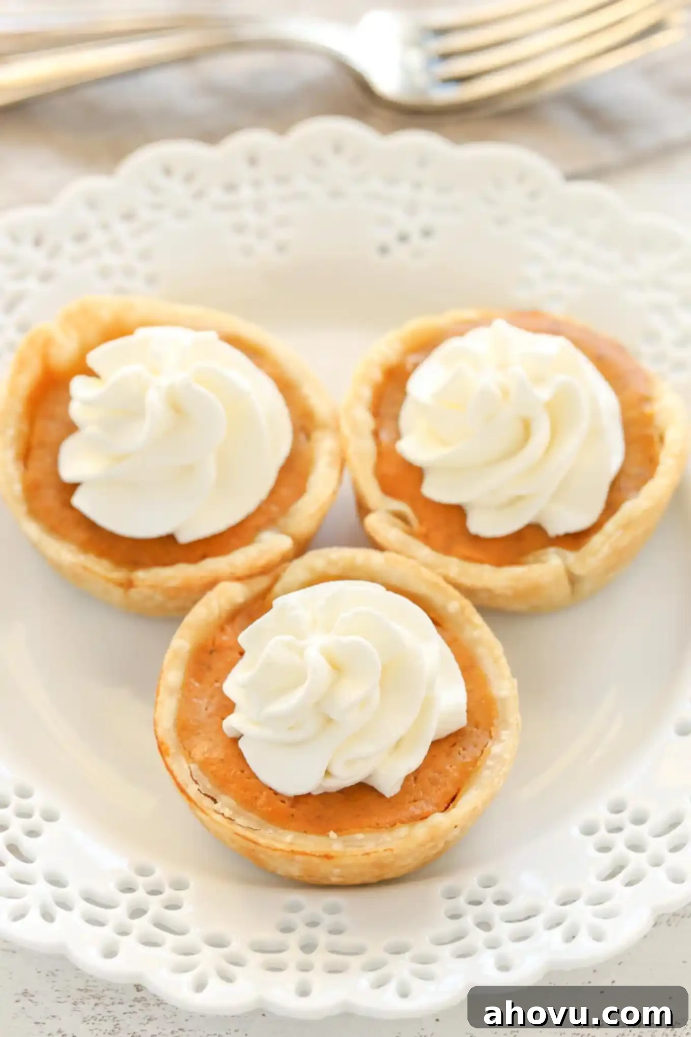 An angled overhead view of three mini pumpkin pies topped with whipped cream on a white plate. Two forks rest in the background.