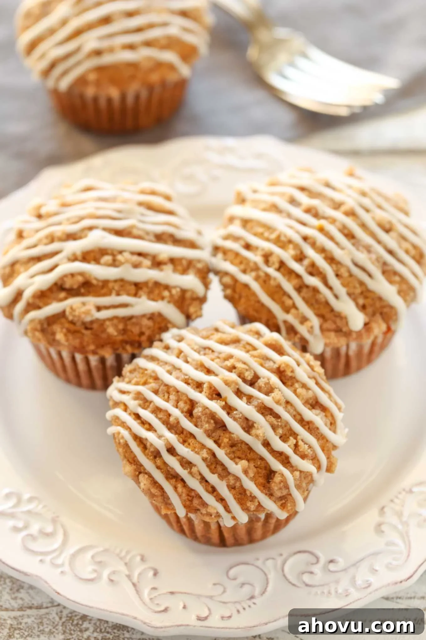 Three glazed pumpkin streusel muffins on a white plate. Another muffin and a fork rest in the background, highlighting the soft interior and golden streusel.
