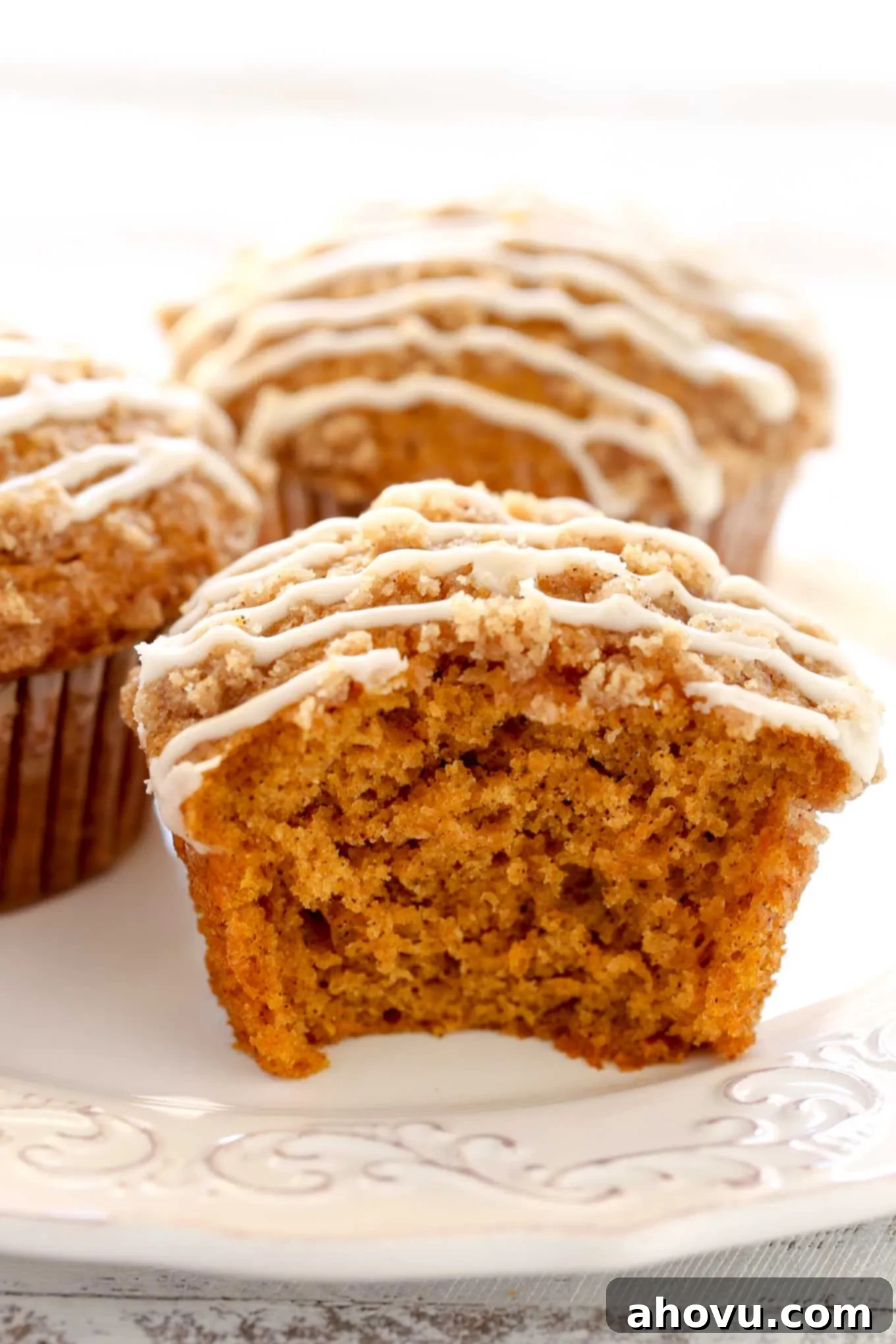 Close up view of pumpkin muffins on a white plate. The front muffin has a bite missing, revealing its moist and tender interior with the distinct crumb topping.