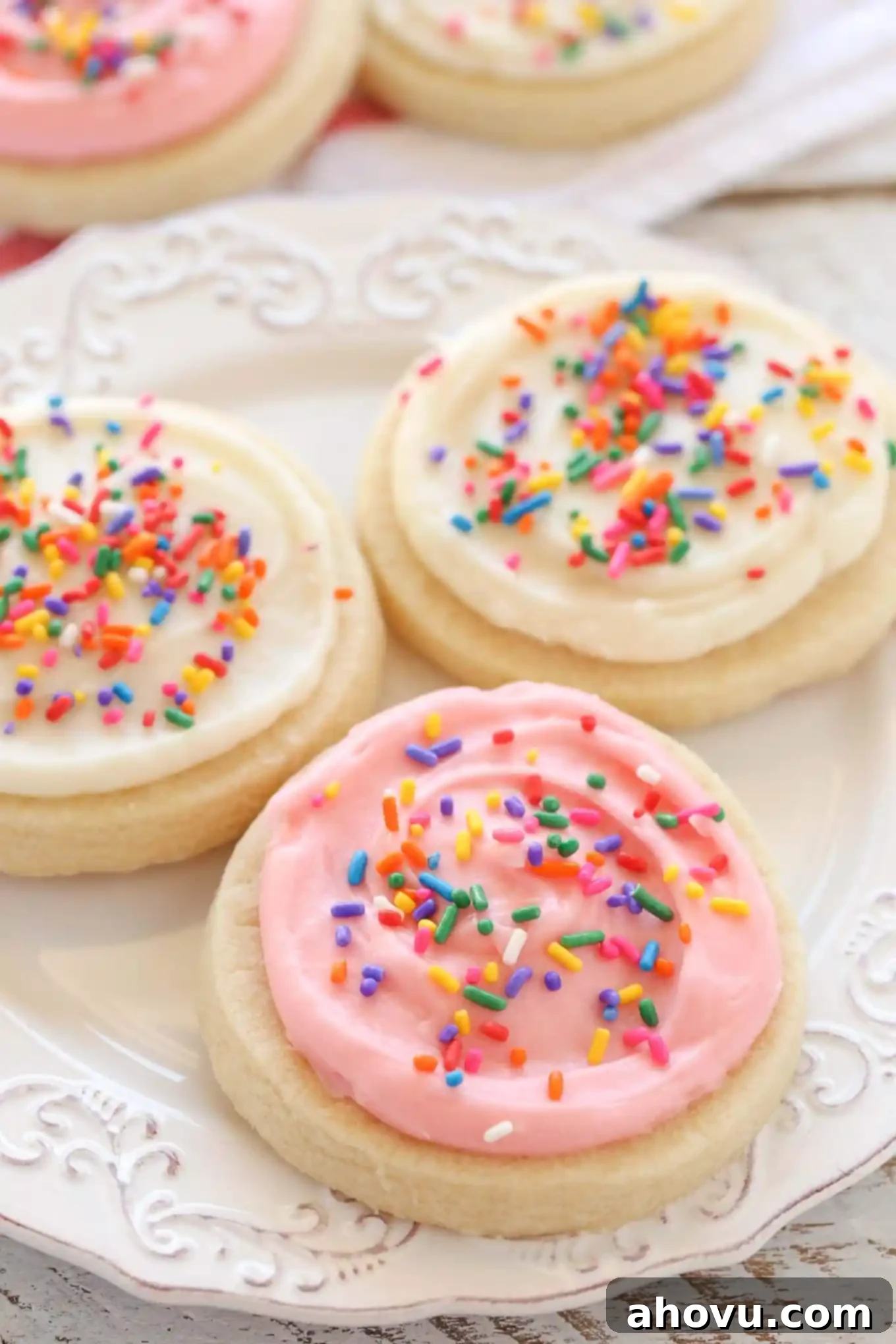 Three frosted sugar cookies on a white plate. Additional sugar cookies rest in the background. 