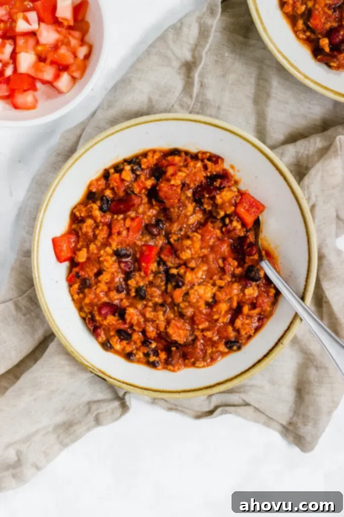 A bowl of turkey chili with a spoon resting in it. Another bowl of diced tomatoes and chili rest nearby.