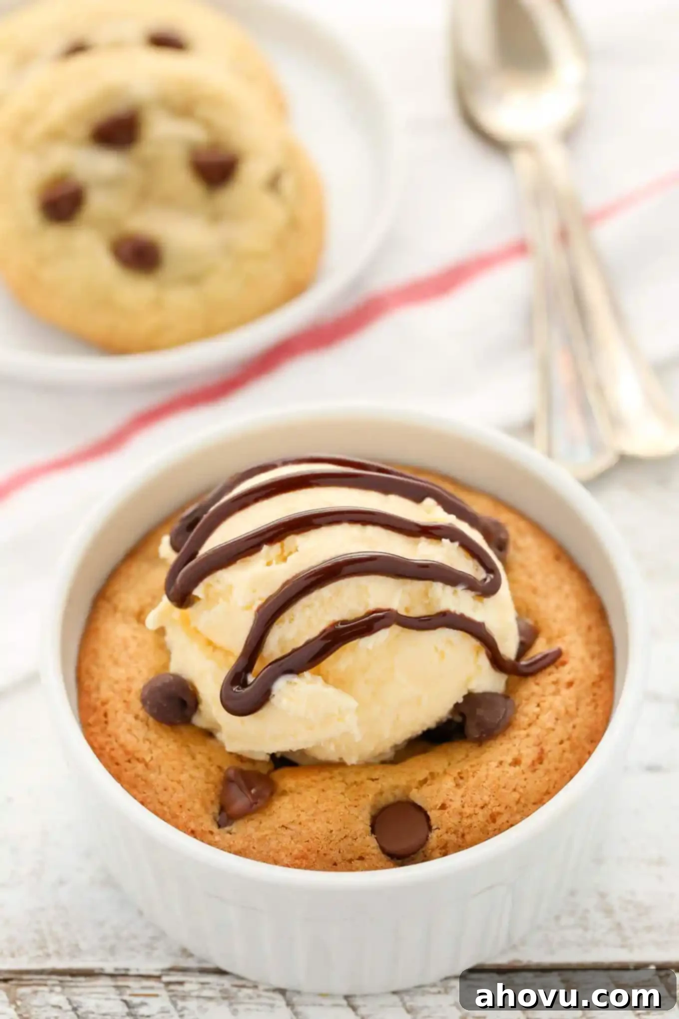 A deep dish single serve chocolate chip cookie topped with vanilla ice cream and chocolate sauce, with a spoon dug in. Two smaller cookies are on a plate in the background.