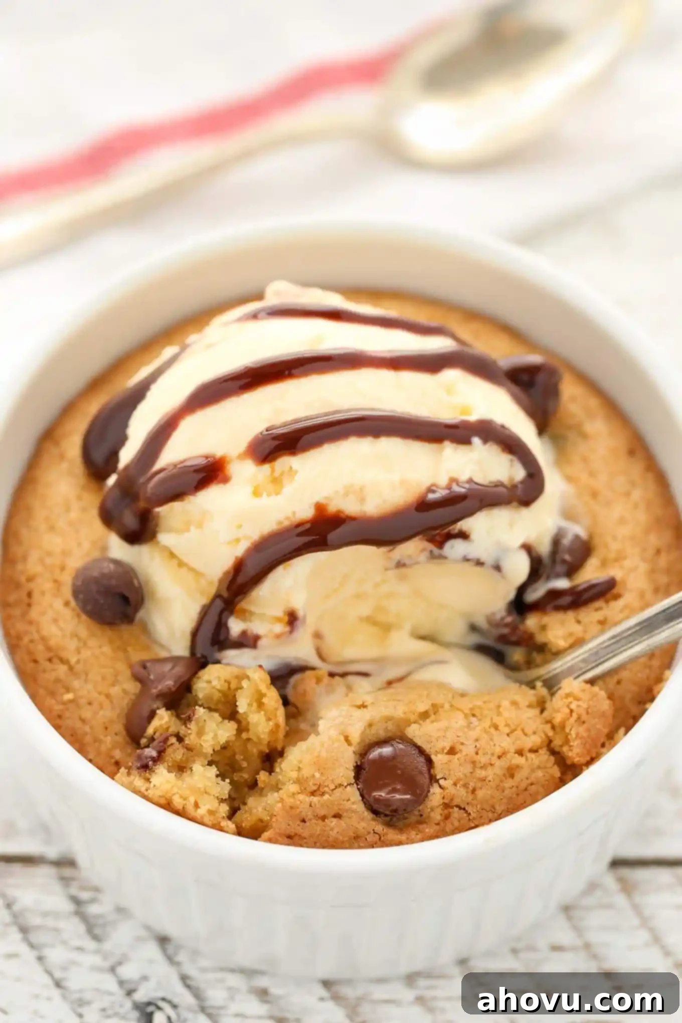 A deep dish single serve chocolate chip cookie topped with ice cream and chocolate sauce, with a spoon being dug into the cookie revealing the gooey center.