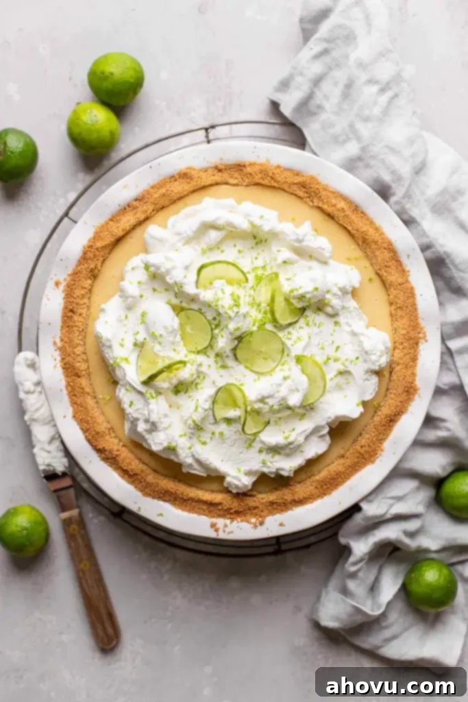 An overhead view of a traditional key lime pie, topped with whipped cream and lime slices. Key limes are scattered around the pie. 