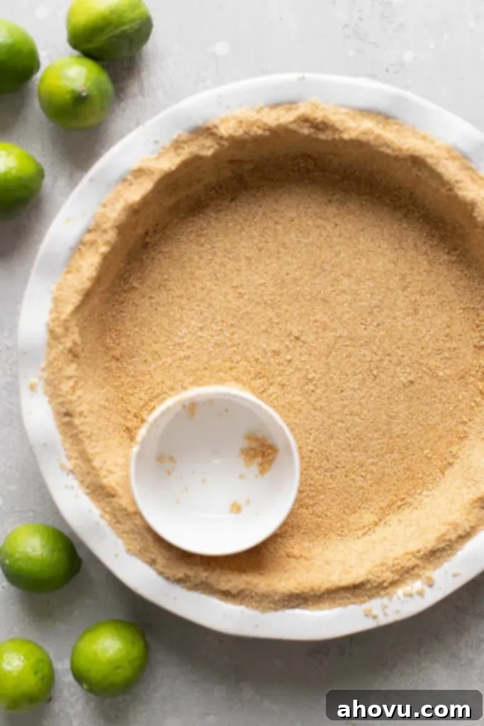 Overhead view of a graham cracker crust in a white pie plate, being packed down with a measuring cup. 