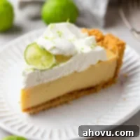 A slice of key lime pie on a white dessert plate. Key limes are scattered in the background.