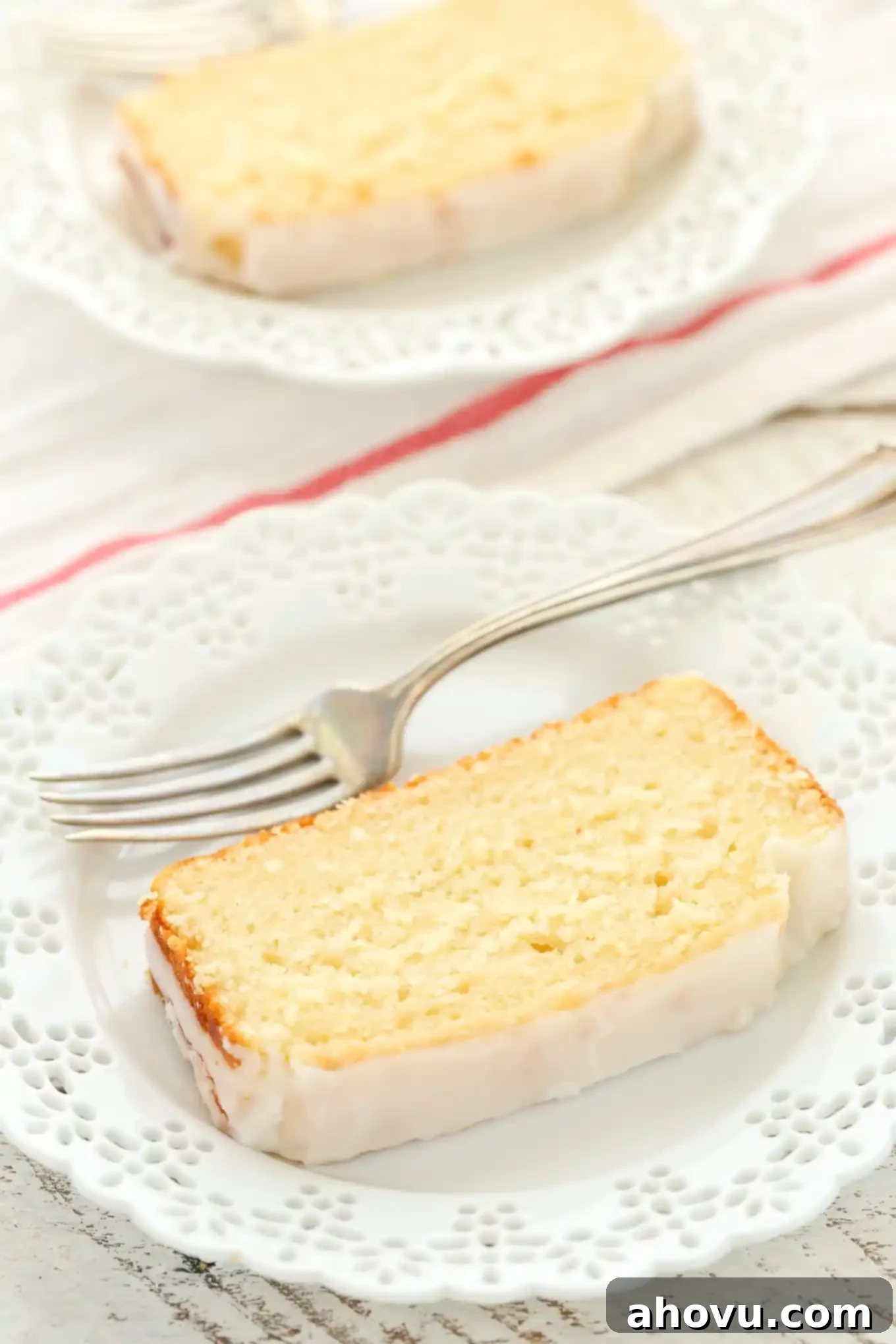 A slice of lemon bread on a white plate with a fork. Another plate rests in the background. 