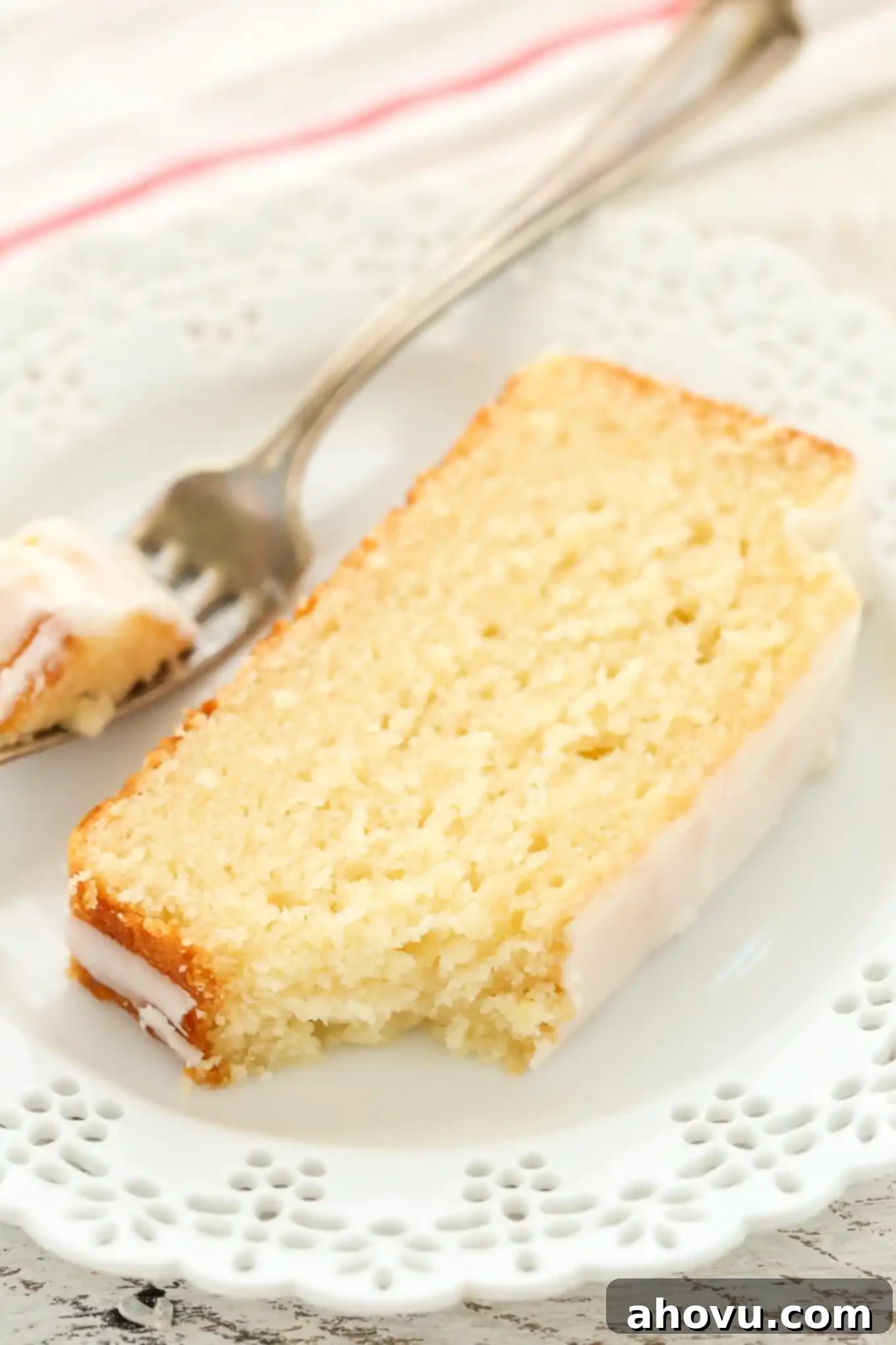 A slice of glazed lemon loaf bread on a plate with a fork. A piece has been removed and is speared on the fork. 