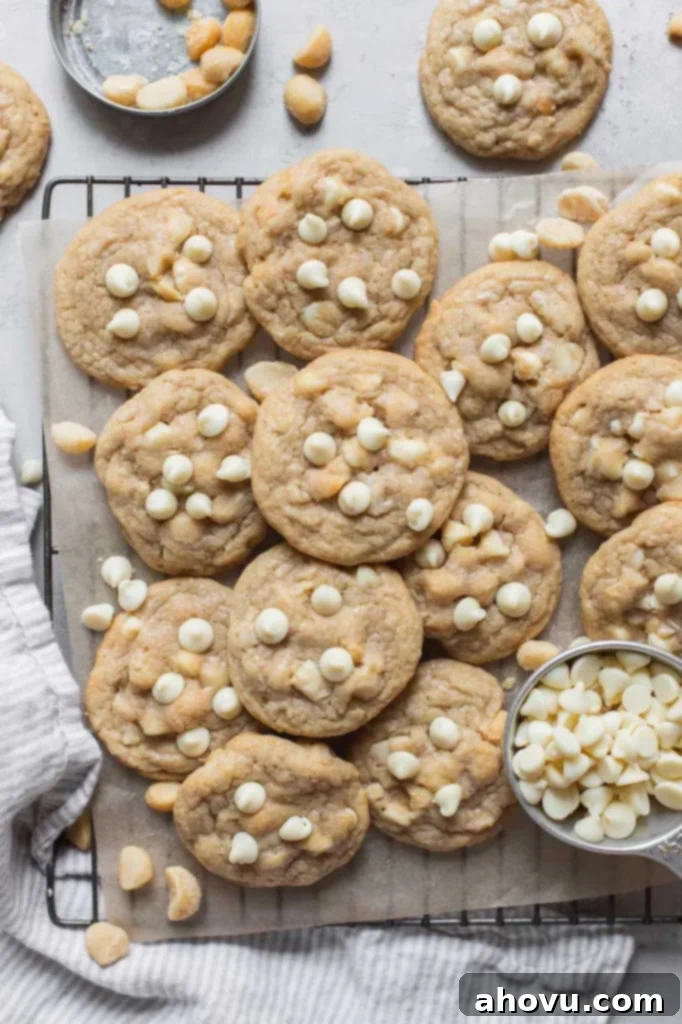 A batch of golden-brown white chocolate macadamia nut cookies laid out on a piece of parchment paper, with extra nuts and white chocolate chips scattered beside them, hinting at the rich inclusions within.
