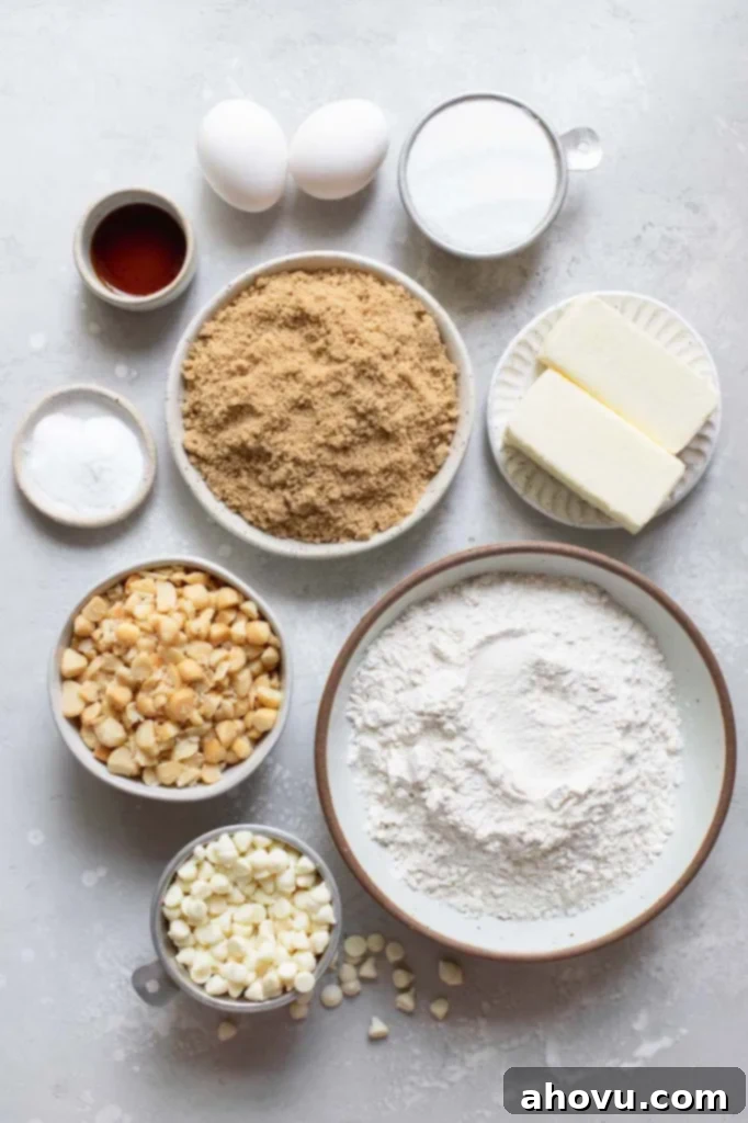 A collection of ingredients neatly arranged in various bowls on a clean gray surface, including flour, sugars, butter, eggs, white chocolate chips, and macadamia nuts, ready for baking.