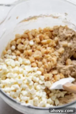 A rubber spatula gently folding white chocolate chips and chopped macadamia nuts into a fluffy cookie dough inside a glass mixing bowl.