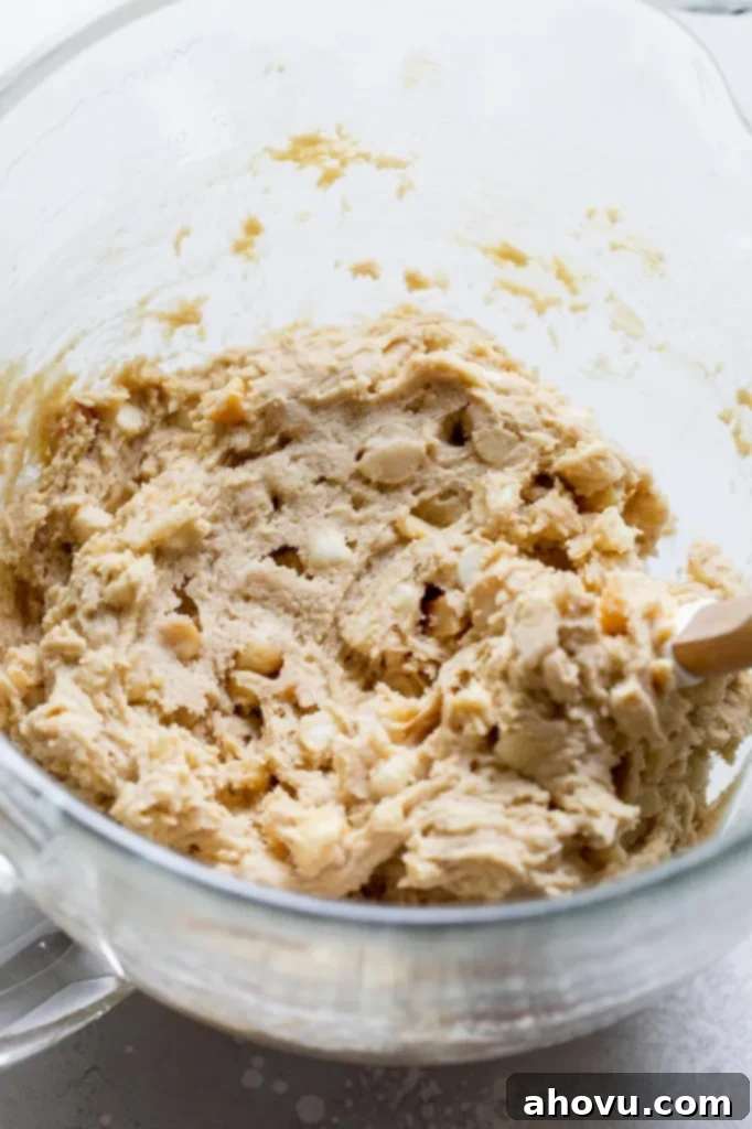 The finished white chocolate macadamia nut cookie dough, rich with chips and nuts, perfectly mixed and ready for chilling in a clear glass mixing bowl.