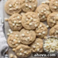 A batch of golden-brown white chocolate macadamia nut cookies laid out on a piece of parchment paper, with extra nuts and white chocolate chips scattered beside them, hinting at the rich inclusions within.