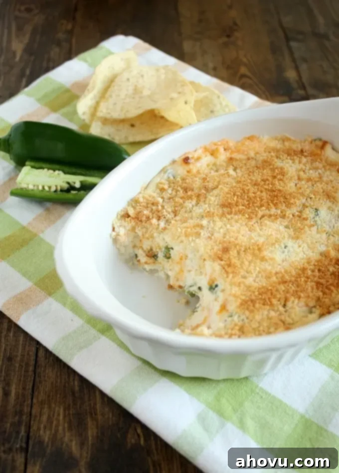 A portion of the creamy jalapeno popper dip has been scooped out, revealing its rich, bubbly texture. It's set in a small baking dish on a checkered towel with fresh jalapenos and tortilla chips nearby.