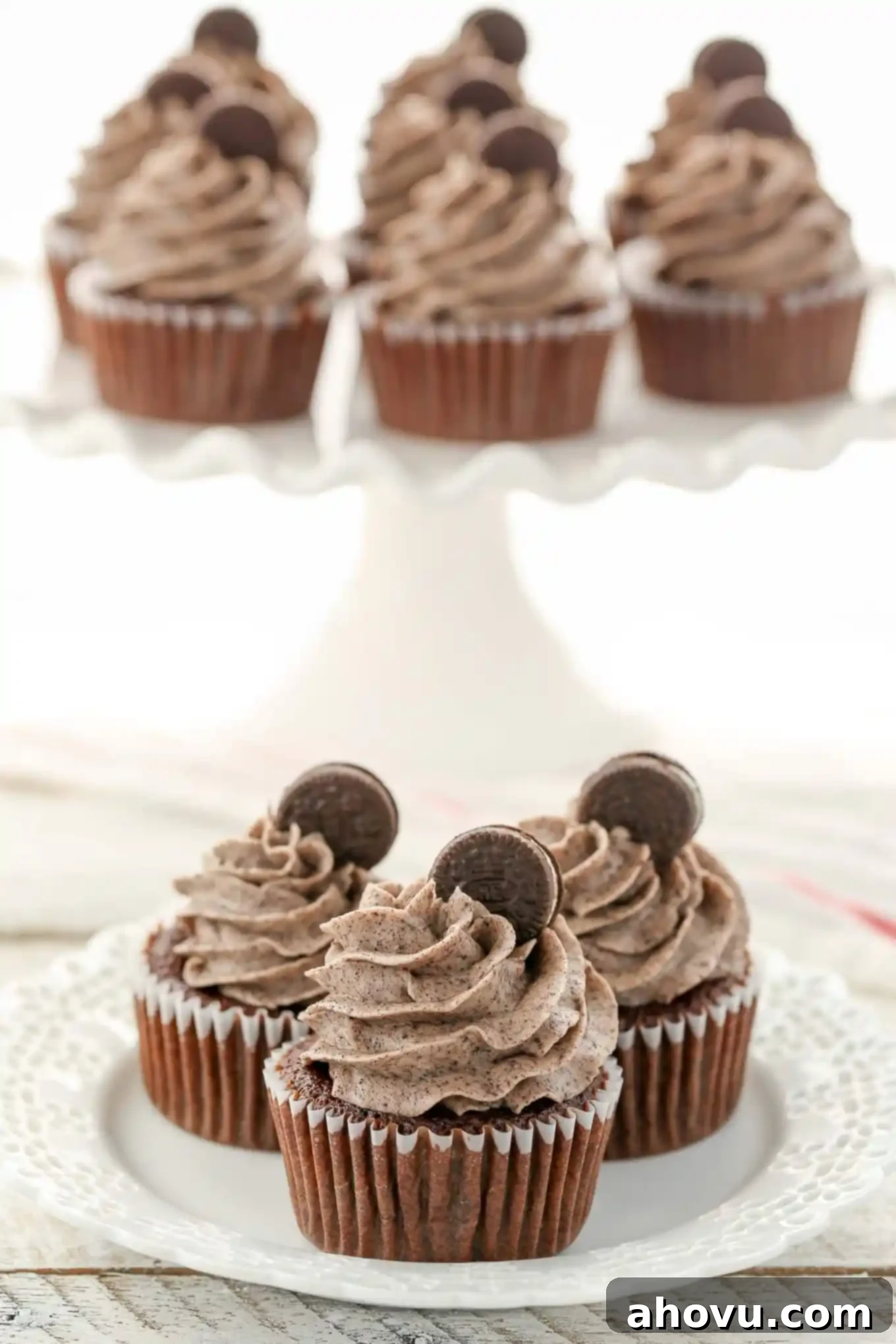 Three oreo cupcakes on a white plate. More cupcakes rest on a cake stand in the background. 