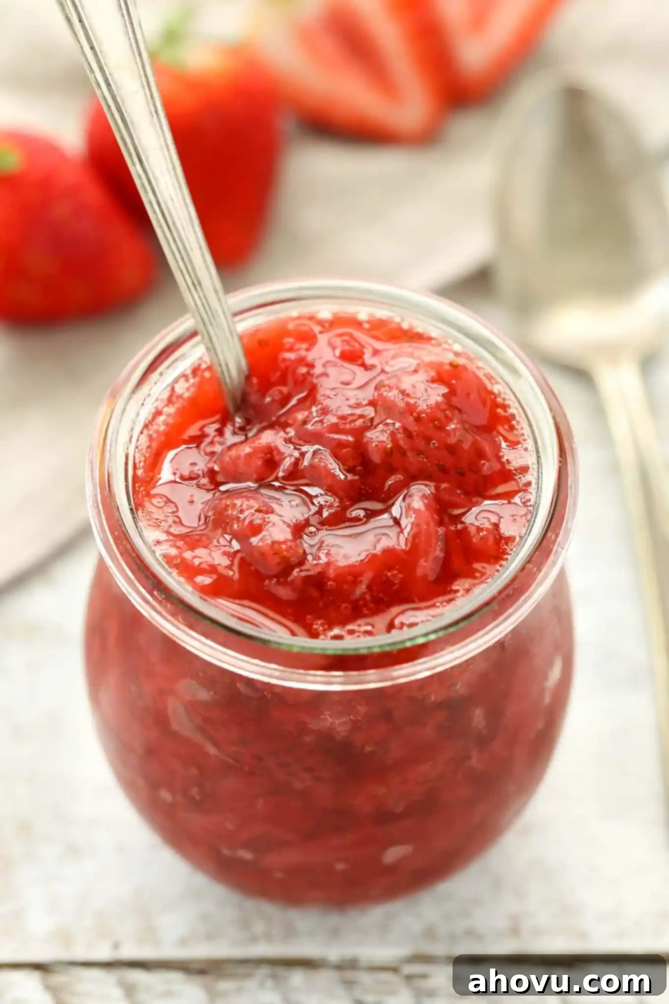 Homemade strawberry sauce in a glass jar with a spoon sticking out. Fresh strawberries rest in the background. 