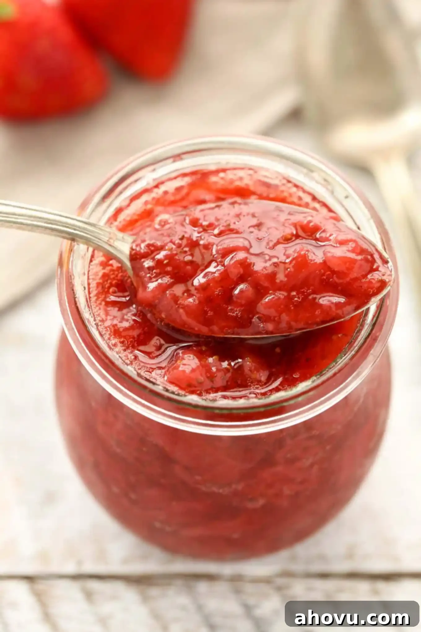 A spoon balanced on top of a glass jar of strawberry sauce. 