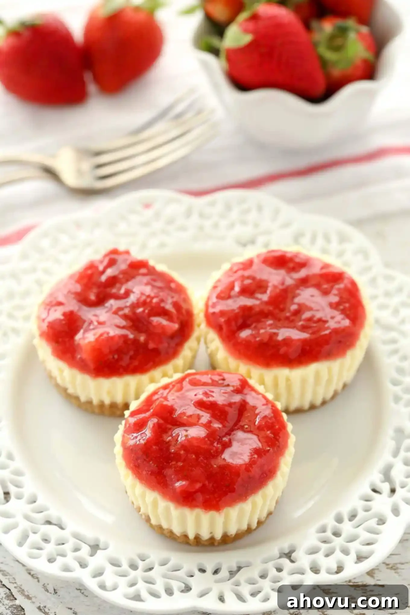 Three homemade strawberry cheesecake bites on a white plate. A fork and a small bowl of fresh strawberries rests in the background. 