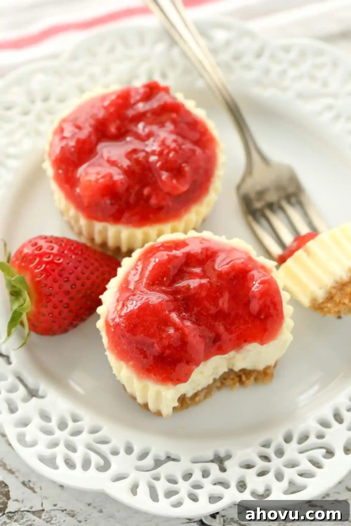 Two strawberry cheesecake bites on a white plate. One mini cheesecake is missing a bite, which is speared on a fork that's also sitting on the plate.