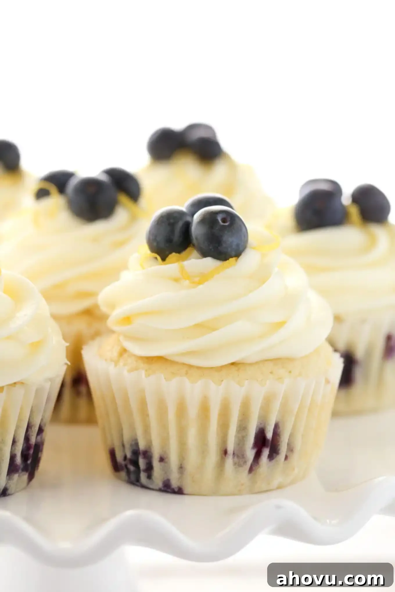 Side view of fluffy lemon blueberry cupcakes topped with swirl of creamy lemon frosting and fresh blueberries on a white cake stand.