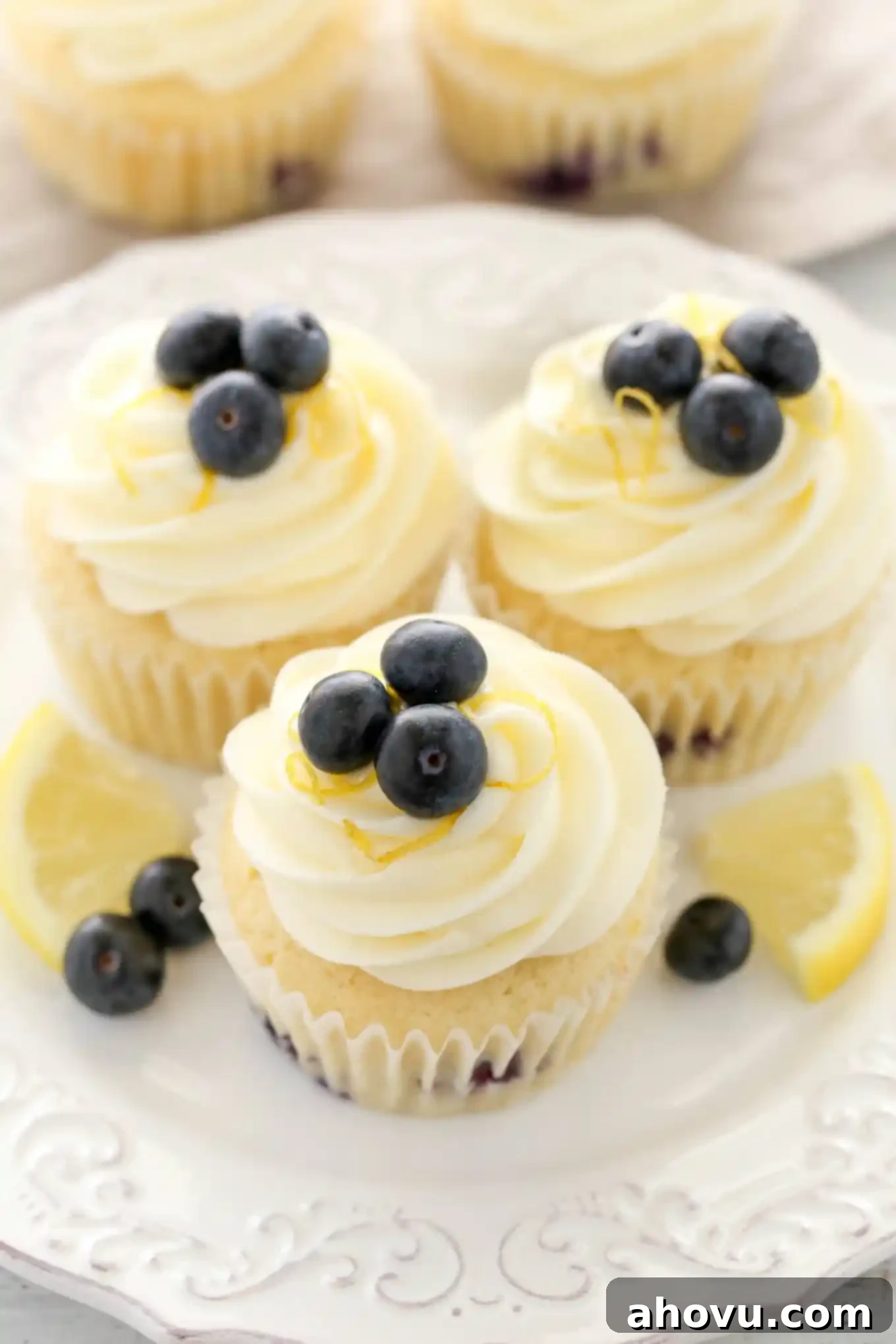Close-up of three beautifully frosted lemon blueberry cupcakes on a white dessert plate, garnished with fresh lemon slices and berries.