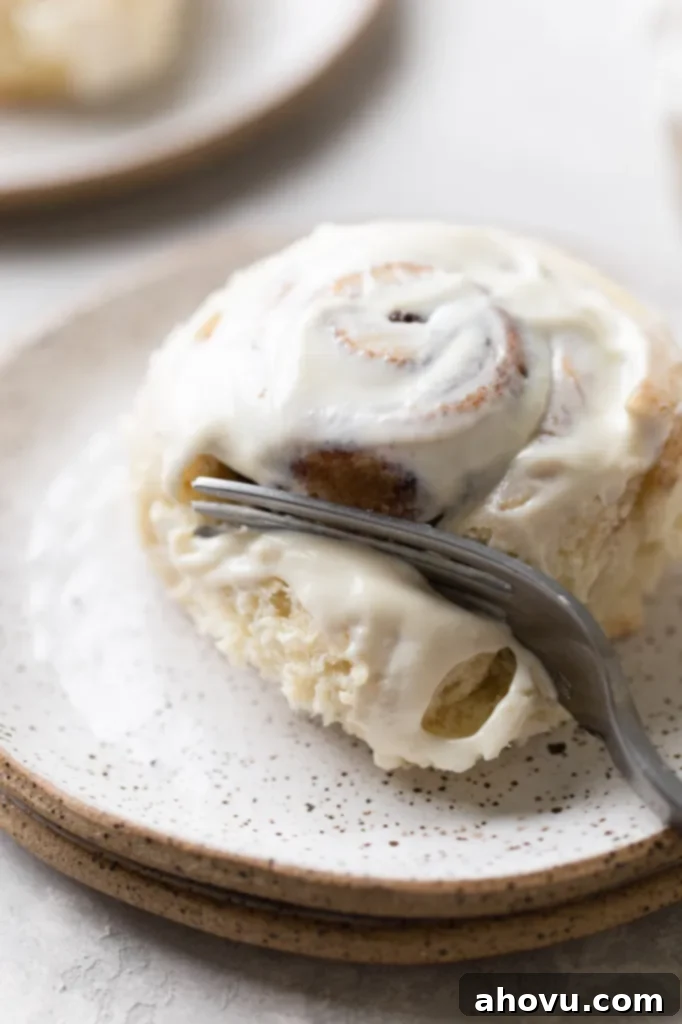 A single, perfectly baked cinnamon roll on a white speckled plate being cut into with a fork, revealing its soft, gooey interior and perfect cinnamon swirls, ready to be enjoyed.