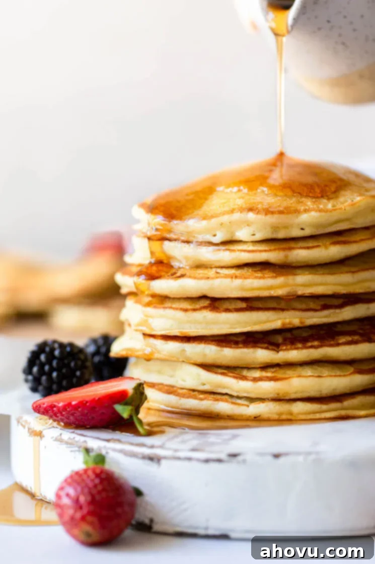 Maple syrup being poured onto a stack of golden-brown buttermilk pancakes on an antique white board, surrounded by fresh, vibrant strawberries and blackberries.