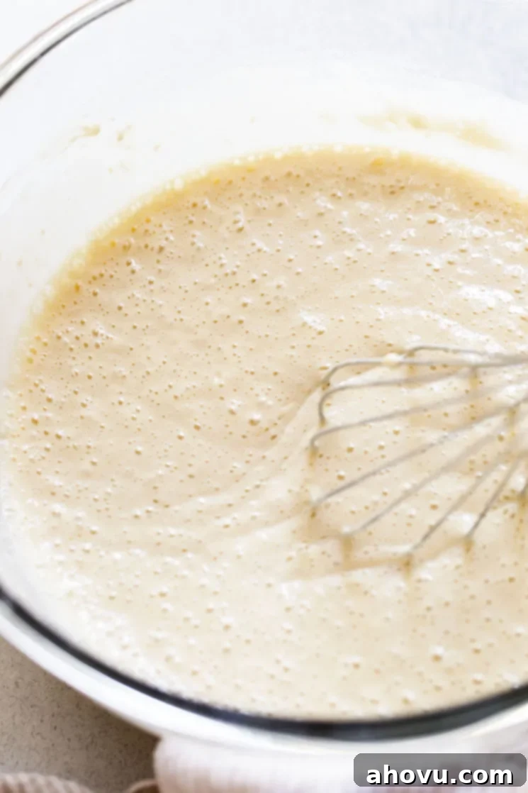 The wet ingredients for buttermilk pancakes, including buttermilk, granulated sugar, oil, vanilla extract, and eggs, being whisked together vigorously in a clear glass mixing bowl, creating a smooth, homogenous mixture.