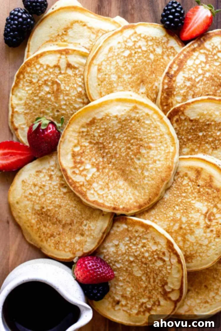 Multiple golden-brown buttermilk pancakes, some stacked and some laying flat, beautifully arranged on a rustic wooden surface, with a side of pure maple syrup and a scattering of fresh mixed berries.