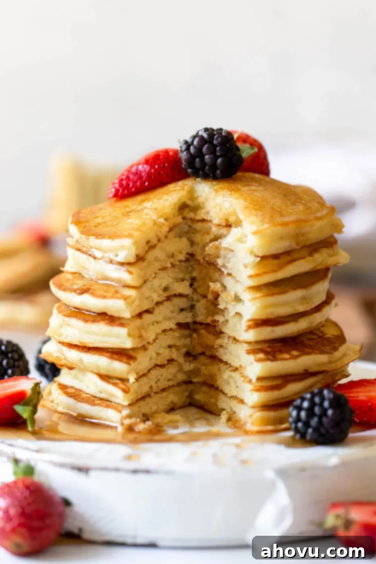 A perfectly stacked and fluffy buttermilk pancake with a bite taken out, revealing its airy texture, topped generously with maple syrup and fresh berries, on an antique white board.