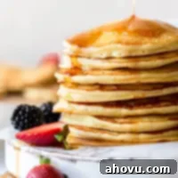 Maple syrup being poured onto a stack of pancakes on an antique white board with strawberries and blackberries surrounding it.