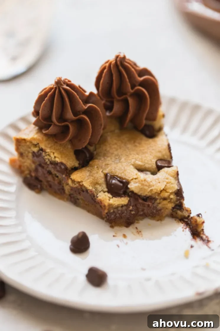 A single, perfectly cut slice of chocolate chip cookie cake, featuring a generous amount of chocolate chips and fluffy buttercream, with a bite taken out of it. It rests on a pristine white plate.