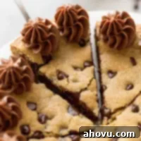 A freshly baked chocolate chip cookie cake sitting on a white cake stand with a slice being removed, showcasing its soft interior and chocolate chips.